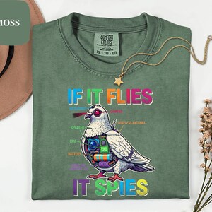 May include: A moss green Comfort Colors t-shirt with the text "IF IT FLIES IT SPIES" and a graphic of a pigeon with tech components. The shirt has a crew neck and a gold star pendant necklace. The shirt is size XL-TG-EG.