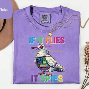 May include: Violet Comfort Colors t-shirt with the text "IF IT FLIES IT SPIES" and a graphic of a pigeon with camera and tech components. A gold star necklace is draped across the shirt.