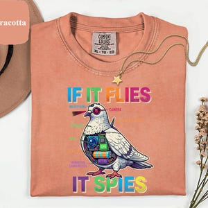 May include: Terracotta-colored t-shirt with the text "IF IT FLIES IT SPIES" and a graphic of a pigeon with spy gadgets. The shirt has a crew neck and a Comfort Colors tag. A gold necklace with a star pendant is draped on the shirt.