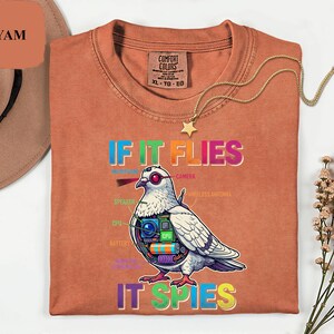 May include: A rust-colored t-shirt with the text "IF IT FLIES IT SPIES" and a graphic of a pigeon with tech components. The shirt has a crew neck and a tag that says "COMFORT COLORS". A gold necklace with a star pendant is also visible.