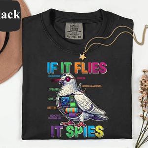 May include: Black t-shirt with the text "IF IT FLIES IT SPIES" and a detailed graphic of a pigeon with tech components. The design includes a microphone, camera, speaker, CPU, battery, and inductive charging coil. A gold necklace with a star pendant is also visible.
