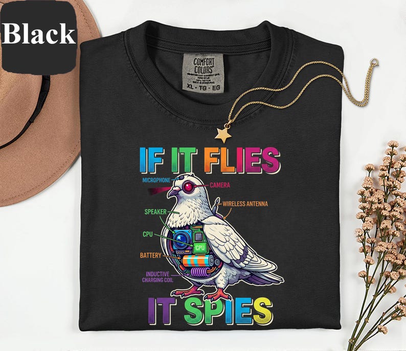 May include: Black t-shirt with the text "IF IT FLIES IT SPIES" and a detailed graphic of a pigeon with tech components. The design includes a microphone, camera, speaker, CPU, battery, and inductive charging coil. A gold necklace with a star pendant is also visible.
