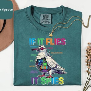 May include: Teal-colored t-shirt with the text "IF IT FLIES IT SPIES" and a graphic of a pigeon with internal tech components. A gold necklace with a star pendant is draped on the shirt.