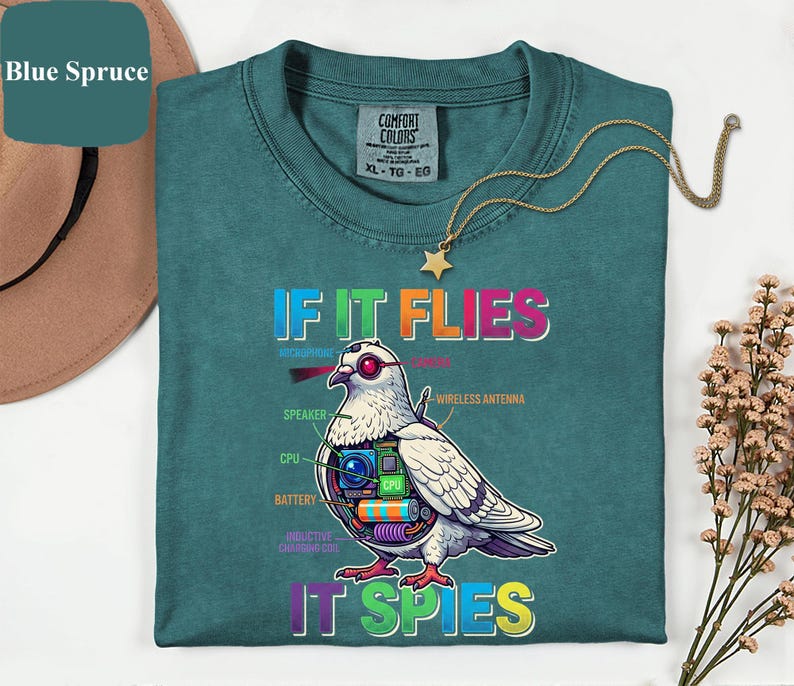 May include: Teal-colored t-shirt with the text "IF IT FLIES IT SPIES" and a graphic of a pigeon with internal tech components. A gold necklace with a star pendant is draped on the shirt.