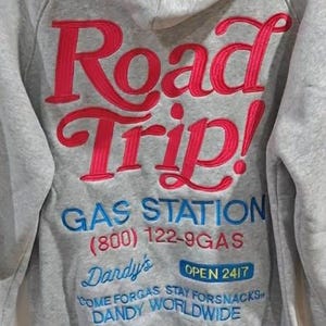 K&ouml;nnte beinhalten: Grauer Kapuzenpullover mit Stickerei. Auf der R&uuml;ckseite steht "Road Trip!" in Rot, dar&uuml;ber "GAS STATION" und weiterer Text in Blau. Der Hoodie hat einen l&auml;ssigen Stil.