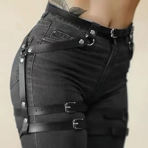 Op de afbeelding: Zwarte leren beenharnas met zilverkleurige gespen en klinknagels. De harnas wordt gedragen over een donkergrijze denim jeans. Het ontwerp omvat riemen die aan een riem zijn bevestigd en de dij omcirkelen.