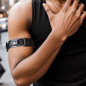 May include: Black leather arm cuff with a silver metal ring. The cuff is worn on the upper arm. The person is wearing a black vest top and a gold chain necklace.