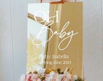May include: Gold mirror sign with white text that reads "Oh Baby" and "Baby Isabella arriving June 2023". The sign is on a wooden easel with pink flowers.