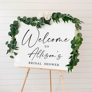 May include: White welcome sign with greenery and white flowers on a wooden easel. The sign reads "Welcome to Allison's Bridal Shower".