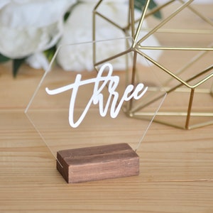 May include: A clear acrylic hexagon table number with the word "three" written in white calligraphy. The table number is on a small wooden base.