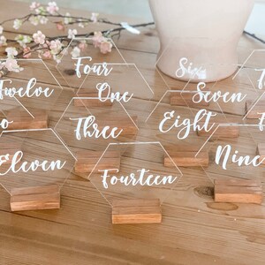 May include: A set of 15 clear acrylic hexagon table numbers with white calligraphy. The numbers are displayed on wooden bases.