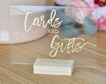 May include: A clear acrylic sign with gold lettering that reads "Cards and Gifts" with a small wooden base that says "Thank You!"