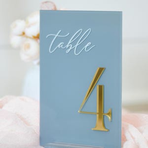 May include: A light blue table number with the word "table" in white script. A gold number 4 is prominently displayed. The table number is on a pink cloth, suggesting a wedding or event theme.