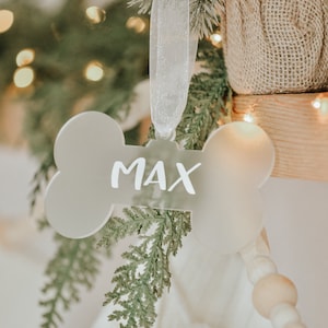 May include: A bone-shaped ornament with the name "MAX" in white lettering. The ornament hangs from a sheer white ribbon, surrounded by evergreen branches and a white stocking. The background has soft, blurred lights.