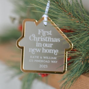 May include: A house-shaped Christmas ornament with gold trim and the text "First Christmas in our new home." The ornament includes the names Katie & William, address, and the year 2023. The ornament hangs from a white ribbon, with pine branches in the background.