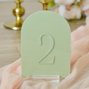 May include: A sage green table number with the number 2 embossed on the front. The arched top table number is displayed on a clear acrylic base. The background includes a pink fabric and gold candle holders.