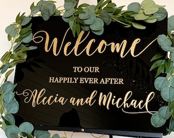 May include: Black welcome sign with gold lettering that reads "Welcome to our happily ever after Alecia and Michael". The sign is decorated with a garland of green leaves.