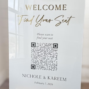 May include: A white welcome sign with gold lettering that reads "WELCOME Find Your Seat." Below the text is a QR code and the names "NICHOLE & KAREEM" with the date "February 7, 2026." The sign is displayed on a wooden easel.