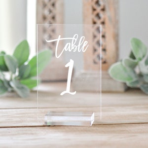 May include: Clear acrylic table number with the word "table" and the number "1" in white script. The table number is rectangular and sits on a small, clear rectangular base. The background is blurred, with wooden surfaces and greenery.