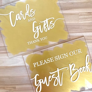 May include: Two acrylic signs with gold paint and white calligraphy. The top sign says "Cards and Gifts Thank You". The bottom sign says "Please Sign Our Guest Book".