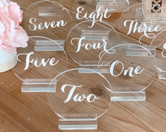 May include: Set of ten clear acrylic table numbers with white calligraphy. The numbers one through ten are displayed on the round acrylic pieces.