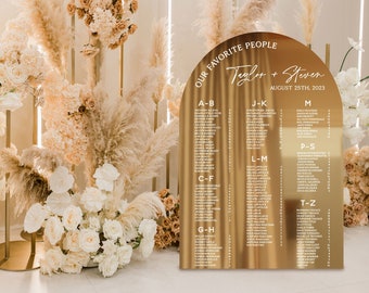 May include: Gold arch-shaped wedding seating chart with the text "Our Favorite People" and "Taylor + Steven" with the date "August 25th, 2023". The chart lists guest names alphabetically by last name.
