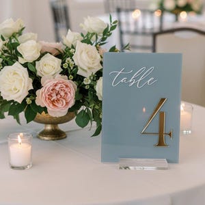 May include: A table setting with a floral arrangement and candles. A blue table number card with gold number 4 and the word "table" in white script. The flowers are white and pink roses in a gold vase.