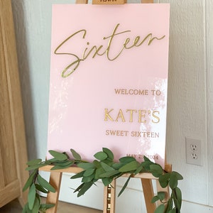 May include: Pink acrylic sign with gold lettering that reads "Sixteen", "Welcome to Kate's Sweet Sixteen", and "May 25, 2024". The sign is on a wooden easel.