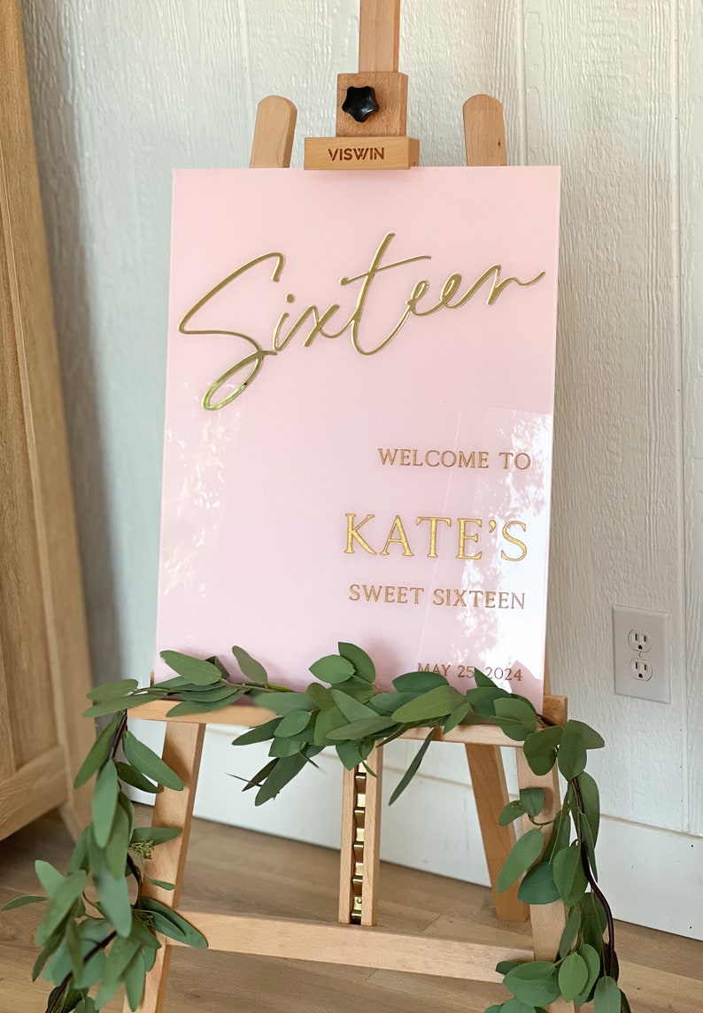 May include: Pink acrylic sign with gold lettering that reads "Sixteen", "Welcome to Kate's Sweet Sixteen", and "May 25, 2024". The sign is on a wooden easel.