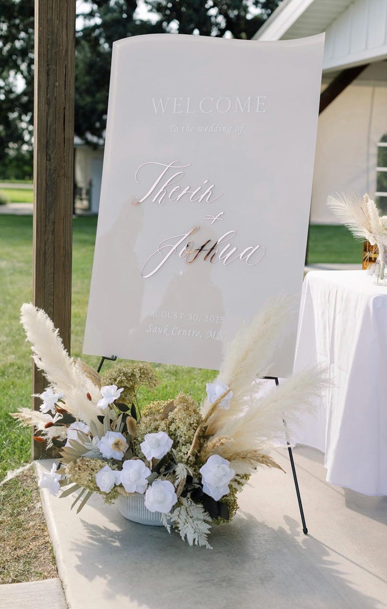 May include: A white wedding welcome sign with the names "Therin" and "Joshua" in elegant script. The sign is propped on a black easel and decorated with white flowers and pampas grass. The date August 30, 2025, and Sauk Centre, MN are also displayed.