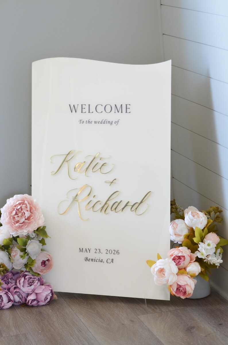 May include: A white welcome sign with gold calligraphy lettering that reads 'Welcome to the wedding of Katie + Richard' with the date 'May 23, 2026' and location 'Benicia, CA' printed below the names.