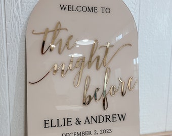 May include: A light pink acrylic sign with a gold script that reads "Welcome to the night before Ellie & Andrew December 2, 2023 Oceanside, CA".