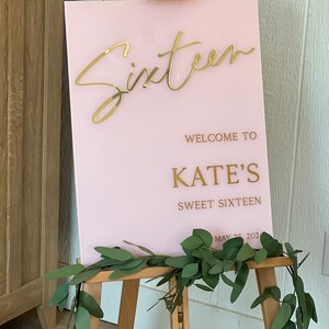 May include: Pink acrylic sign with gold lettering that reads "Sixteen" and "Welcome to Kate's Sweet Sixteen May 25, 202.