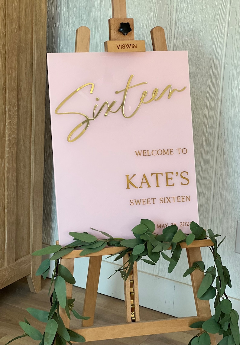 May include: Pink acrylic sign with gold lettering that reads "Sixteen" and "Welcome to Kate's Sweet Sixteen May 25, 202.