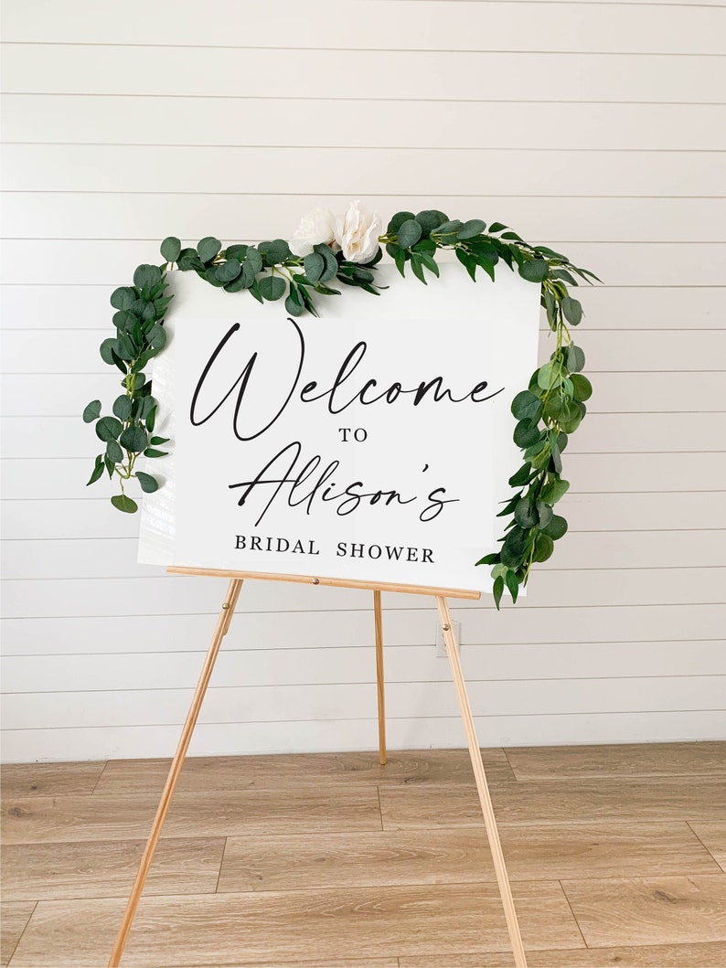 May include: White welcome sign with greenery and white flowers on a wooden easel. The sign reads "Welcome to Allison's Bridal Shower".
