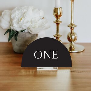 May include: Black and white table number with the word "ONE" in white text on a black half-circle background. The table number is on a clear acrylic stand.