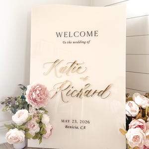 May include: A cream-colored wedding welcome sign with gold lettering that reads "Welcome to the wedding of Katie & Richard" and the date May 23, 2026, in Benicia, CA. The sign is decorated with pink and white flowers.