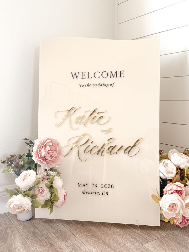 May include: A cream-colored wedding welcome sign with gold lettering that reads "Welcome to the wedding of Katie & Richard" and the date May 23, 2026, in Benicia, CA. The sign is decorated with pink and white flowers.