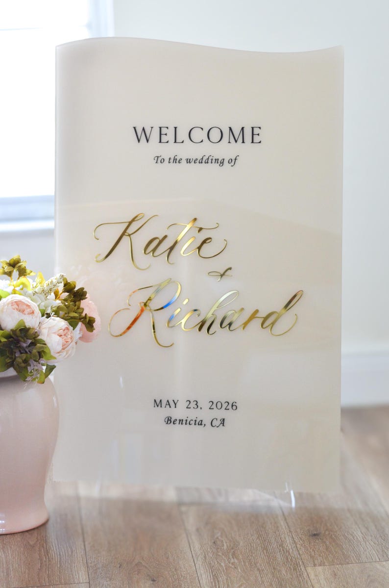 May include: A white welcome sign with gold lettering. The sign reads 'Welcome to the wedding of Katie & Richard, May 23, 2026, Benicia, CA.'