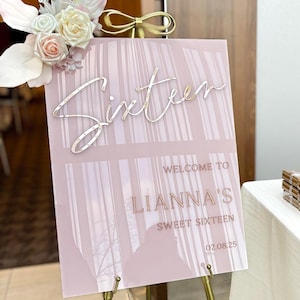 May include: A pink acrylic sign with a white and pink floral design on top. The sign says "Sixteen" in cursive script, "Welcome to LIANNA'S Sweet Sixteen" and "02.08.25".