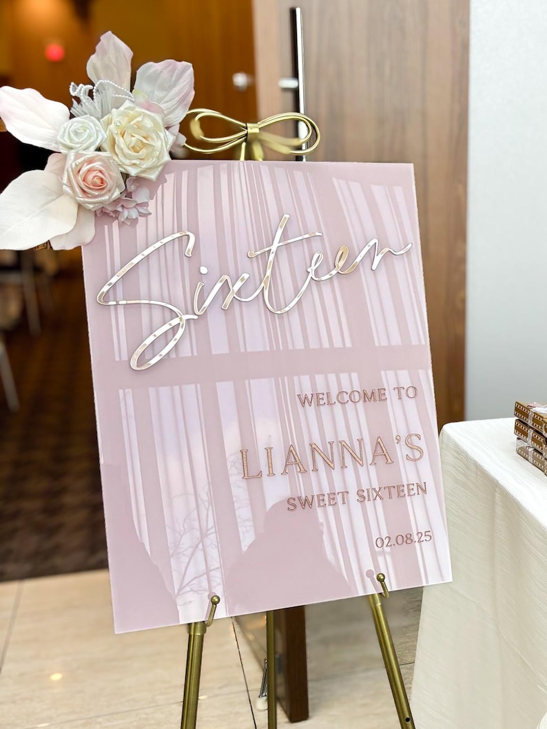 May include: A pink acrylic sign with a white and pink floral design on top. The sign says "Sixteen" in cursive script, "Welcome to LIANNA'S Sweet Sixteen" and "02.08.25".