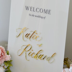 May include: A white wedding welcome sign with gold lettering that reads "WELCOME" and "To the wedding of Katie & Richard." The date is May 23, 2026, in Benicia, CA. Pink and white flowers are in the corner.