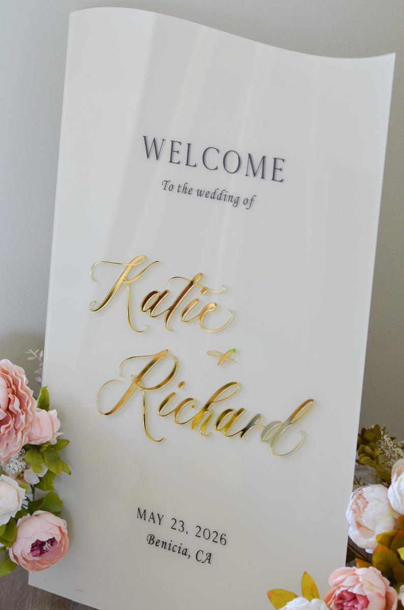 May include: A white wedding welcome sign with gold lettering that reads "WELCOME" and "To the wedding of Katie & Richard." The date is May 23, 2026, in Benicia, CA. Pink and white flowers are in the corner.