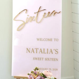 May include: A pink acrylic sign with gold lettering that reads "Sixteen" and "Welcome to Natalia's Sweet Sixteen August 23, 2025".
