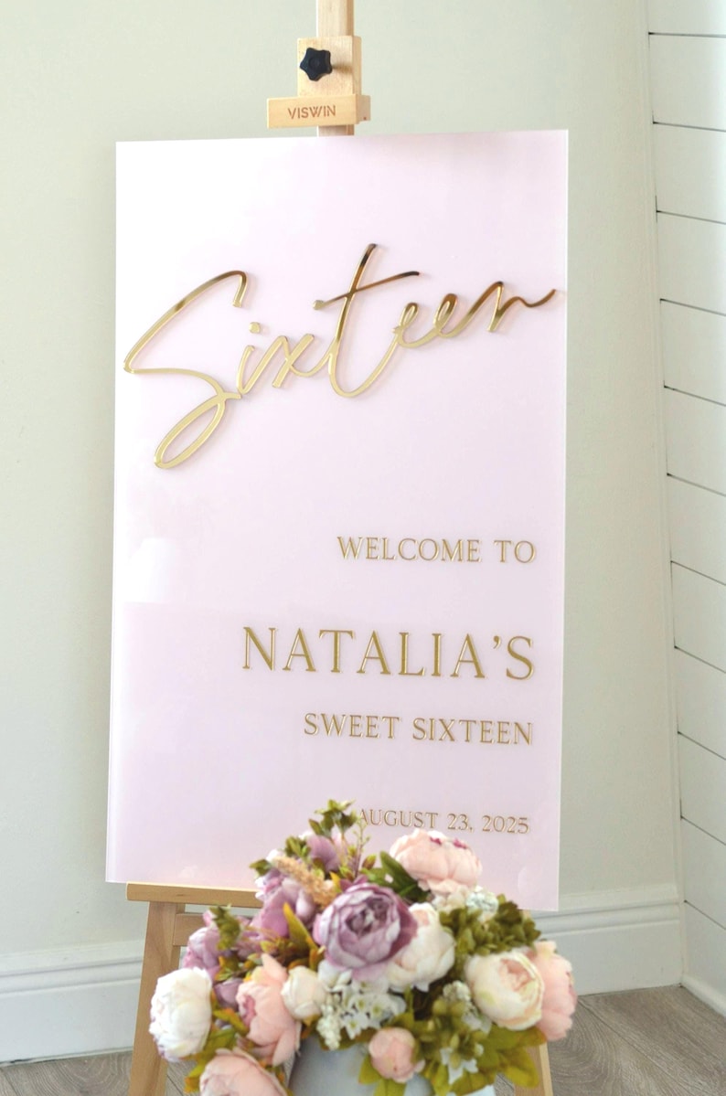 May include: A pink acrylic sign with gold lettering that reads "Sixteen" and "Welcome to Natalia's Sweet Sixteen August 23, 2025".