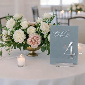 May include: A table setting with a floral arrangement of white and pink roses in a gold vase. A blue-gray table number 4 with silver accents and the word "table" in script is displayed on the table. Lit candles add ambiance.
