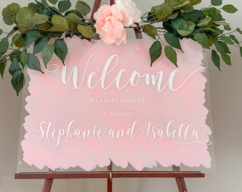 Puede incluir: Bienvenido a un baby shower en honor a Stephanie e Isabella. Un letrero de acrílico rosa y blanco con caligrafía blanca en un caballete de madera con vegetación y flores.