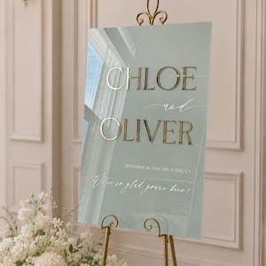May include: A rectangular sage green acrylic wedding sign featuring raised gold lettering that reads CHLOE and OLIVER, with the date SEPTEMBER 26, 2026, location HILLS SIDE, CA, and the script text We're so glad you're here! displayed on a gold easel.