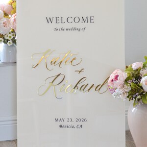 May include: A cream-colored wedding welcome sign with gold lettering. The sign reads "WELCOME To the wedding of Katie + Richard MAY 23, 2026 Benicia, CA." Flowers in vases are on either side of the sign.