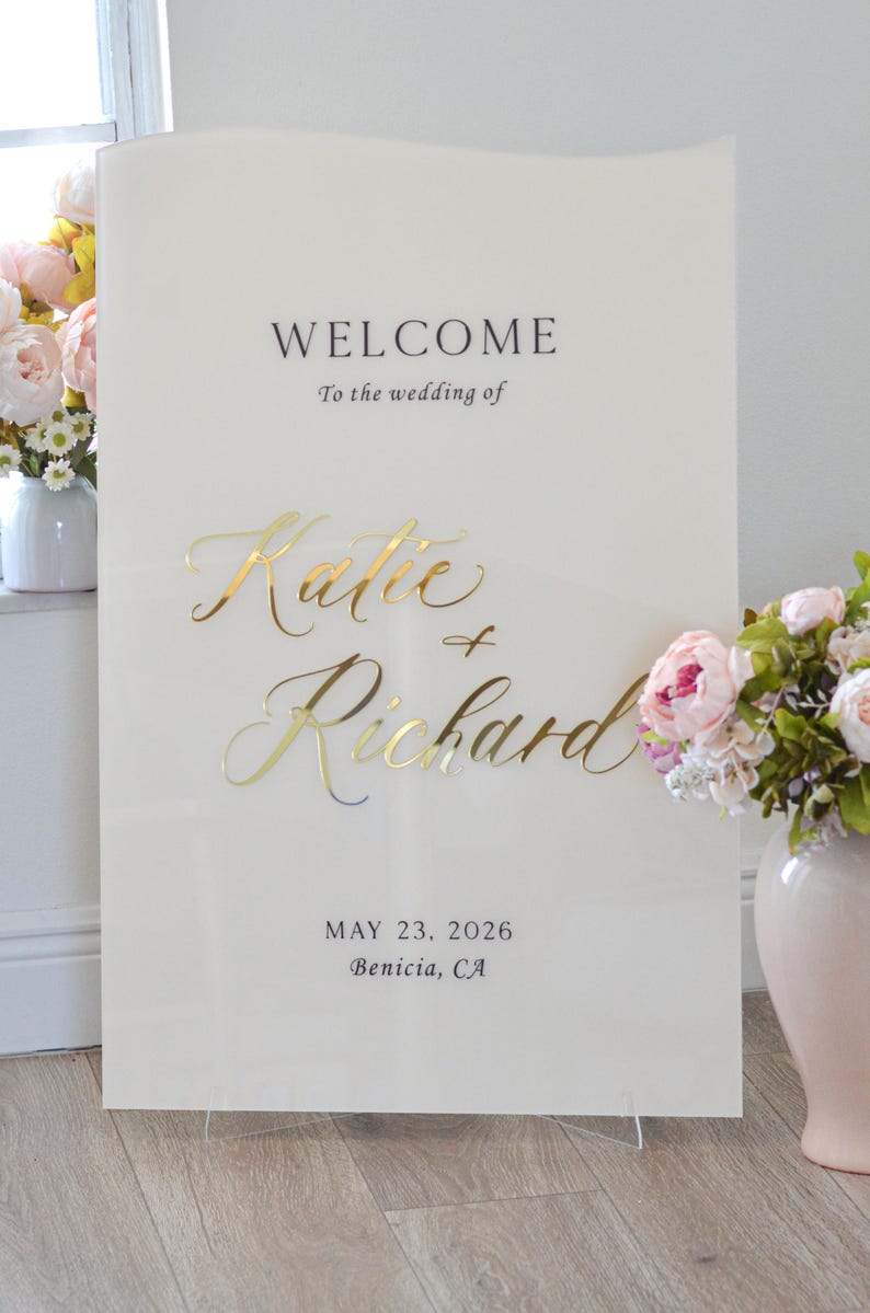May include: A cream-colored wedding welcome sign with gold lettering. The sign reads "WELCOME To the wedding of Katie + Richard MAY 23, 2026 Benicia, CA." Flowers in vases are on either side of the sign.